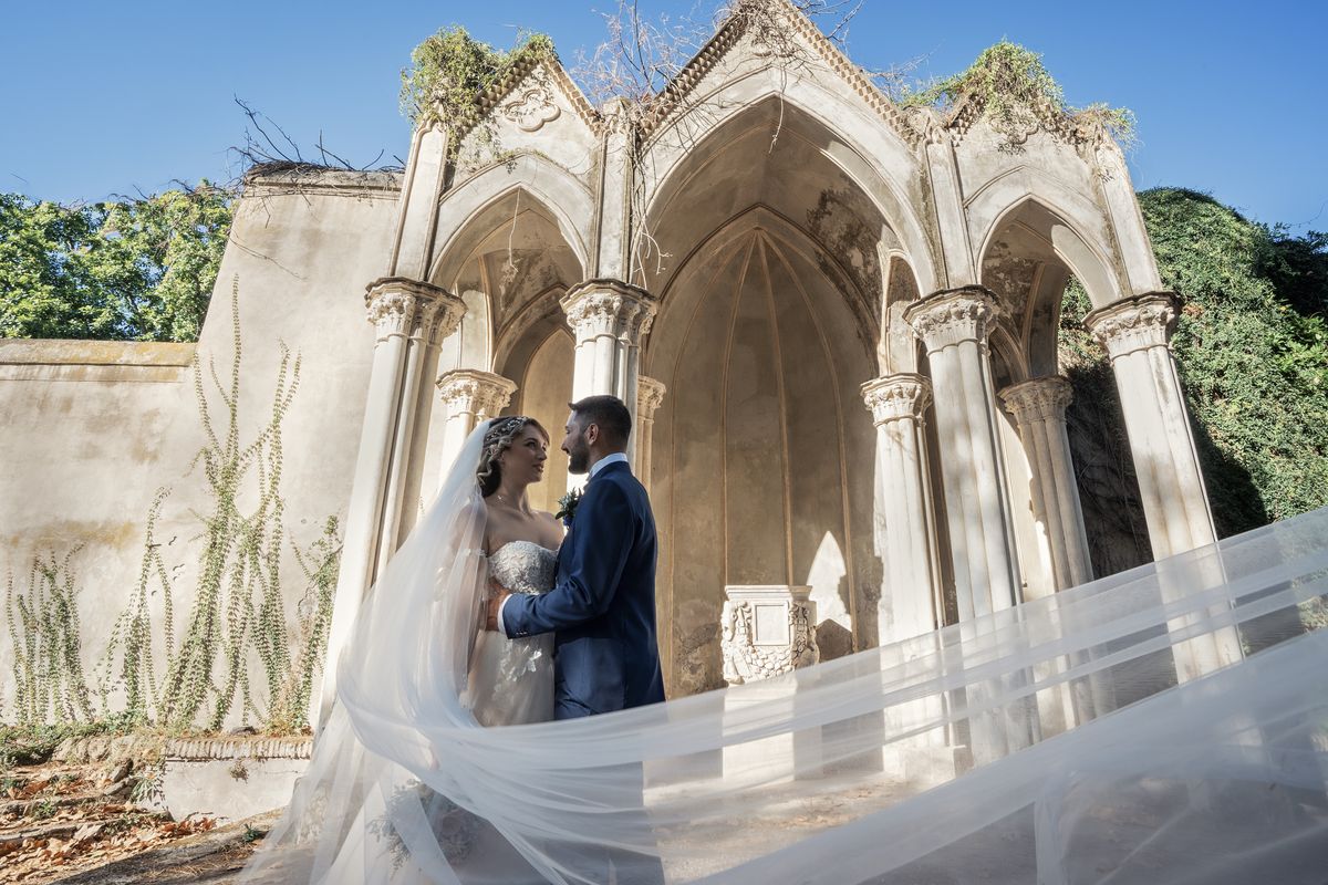 Sposi con velo al vento sotto gli archi gotici di un giardino storico romano